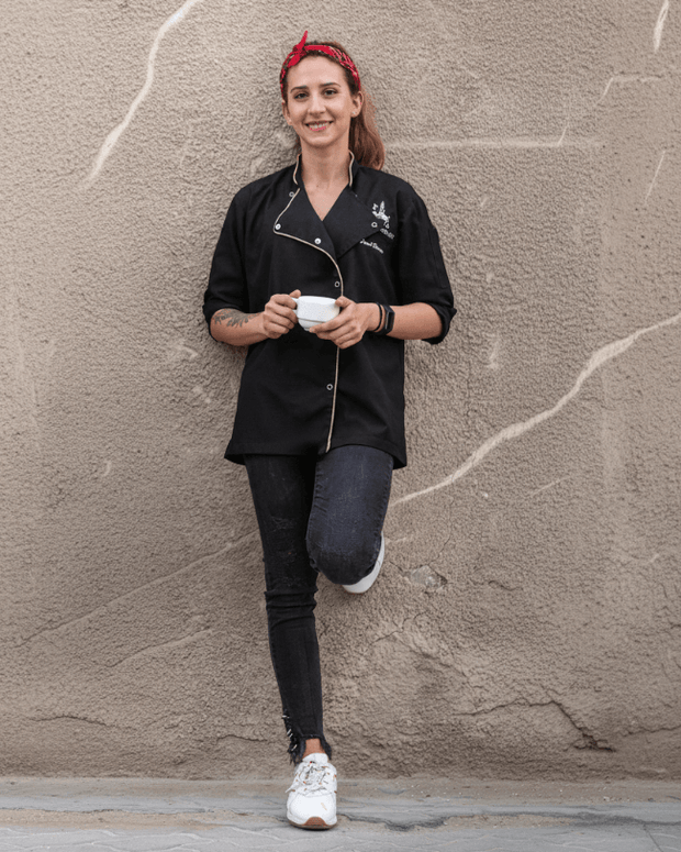 Farah Hassan, co-founder, chef patissier of Flour Girl Bakeries UAE Dubai Abu Dhabi, wearing a black apron with Flour Girl logo, holding a white cup against a textured wall.