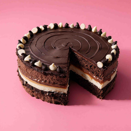 Flour Girl's Dalmation Cake, a chocolate cake with a slice removed, showing layers of dark chocolate cake, caramel sauce, and a cream-colored filling, topped with chocolate and white chocolate chips, set against a solid pink background.
