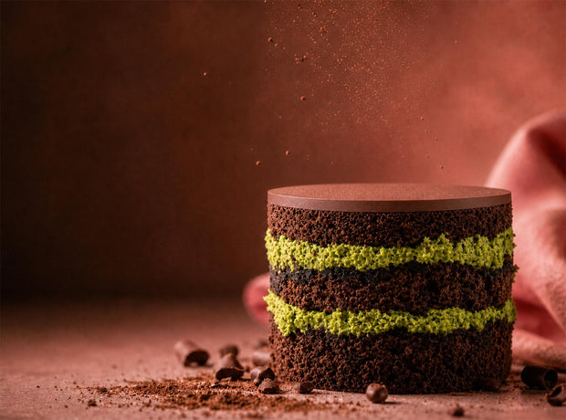 A pistachio kunafa chocolate mono cake with pistachio kunafa spread and chocolate sponge cake layers, photographed on a warm brown surface with cocoa powder and chocolate chips scattered around it.
